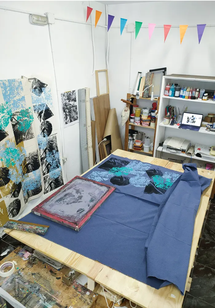 Imagen de parte del taller donde se muesta diferentes diseños expuestos y una tela azul sobre una mesa con estampaciones y una pantalla de impresión, al fondo diferentes colores