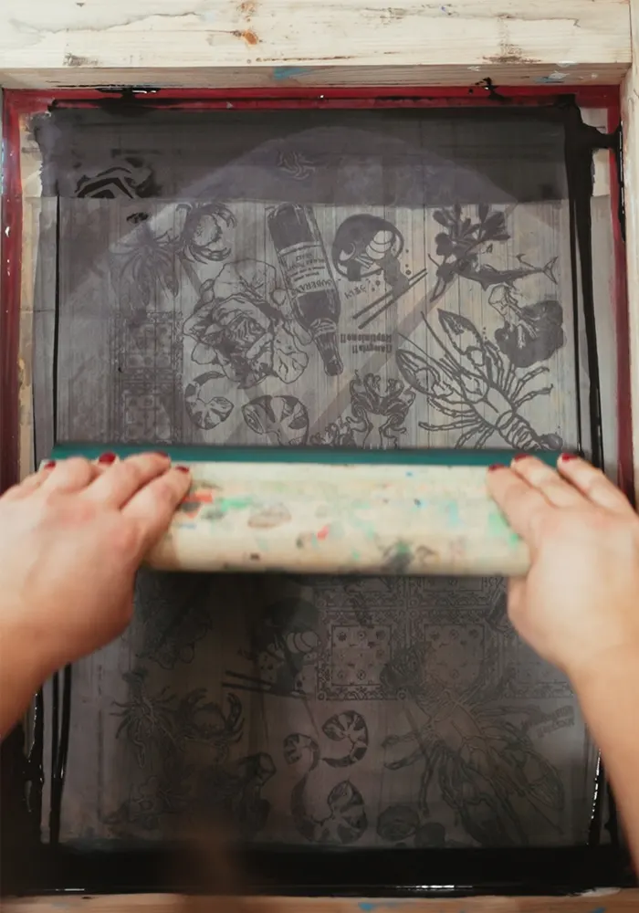 Detalle de la impresión con serigrafía de un diseño, se muestan dos manos de mujer moviendo la rasqueta para realizar la impresión