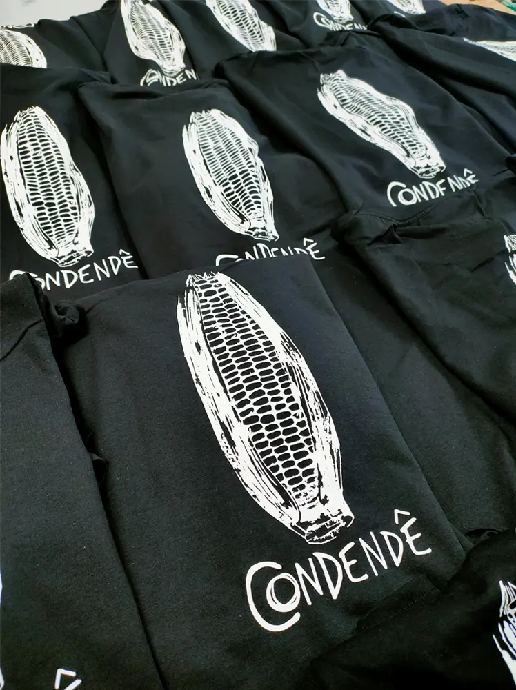 Varias camisetas negras expuestas con un diseño en color blanco de Condendé y una mazorca de maiz impresa en blanco