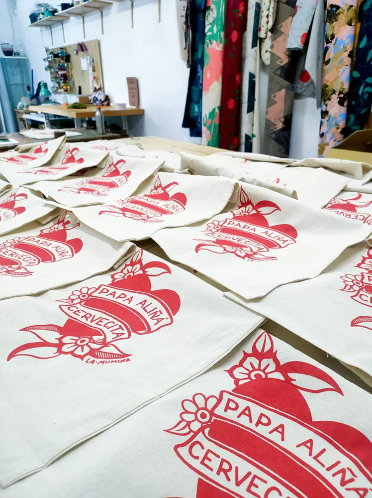 Totebags expuestas sobre una mesa de madera con un diseño de un corazón en rojo con la frase ""Papa aliña cervecita" de La Mumina
