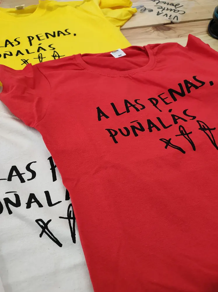 Camisetas de diferentes colores básicos estampadas con un diseño que pone " a las penas, puñalás" seguido de la la imagen de 3 puñales en color negro